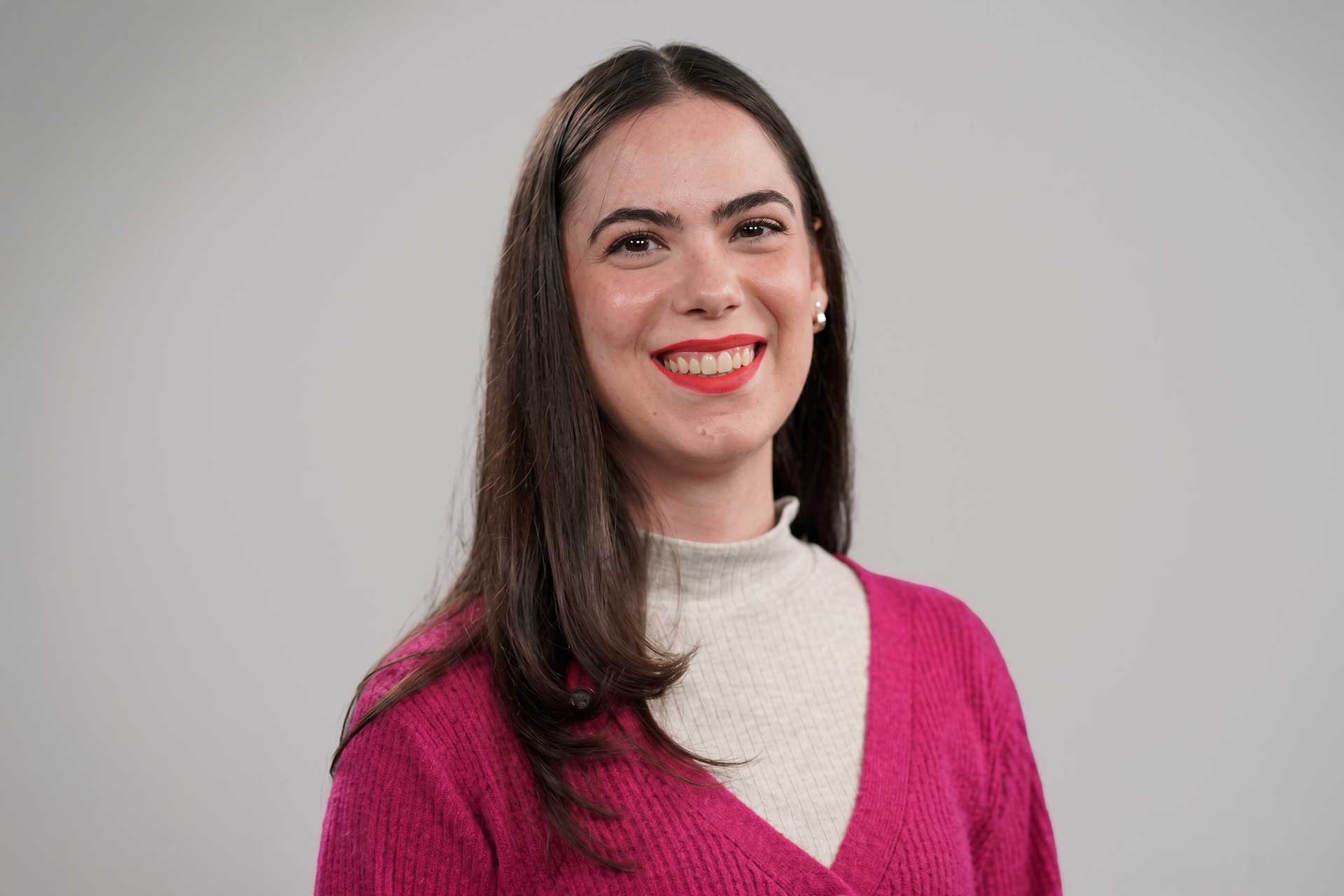 Retrato da jornalista Jamille Niero sorrindo, vestindo cardigan rosa e blusa off-white, contra um fundo cinza.