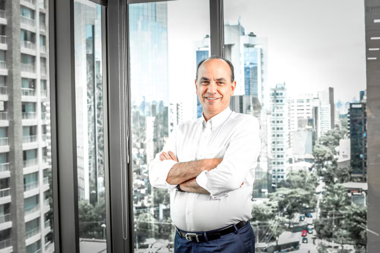 Homem de camisa branca e calça azul posa de braços cruzados diante de uma janela com vista para prédios comerciais. A imagem mostra Fernando Simões, CEO da Simpar, em um escritório.