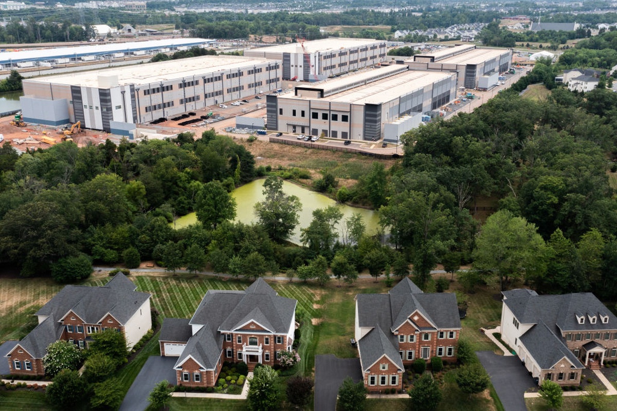 Vista aérea de um grande complexo de data centers cercado por vegetação e localizado próximo a um bairro residencial. Em primeiro plano aparecem casas unifamiliares; ao fundo, os galpões industriais que abrigam a infraestrutura de processamento de dados.