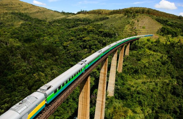 Trem de passageiros atravessa uma ponte ferroviária elevada sobre uma área de mata e morros verdes, em região montanhosa do Brasil. A composição faz uma curva em meio à paisagem rural.