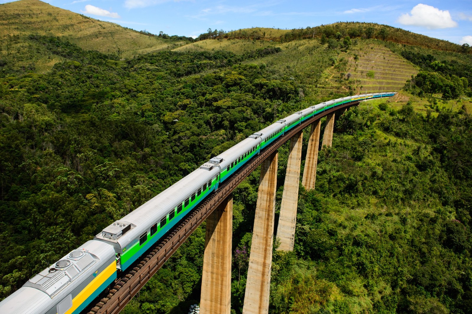 Trem de passageiros atravessa uma ponte ferroviária elevada sobre uma área de mata e morros verdes, em região montanhosa do Brasil. A composição faz uma curva em meio à paisagem rural.