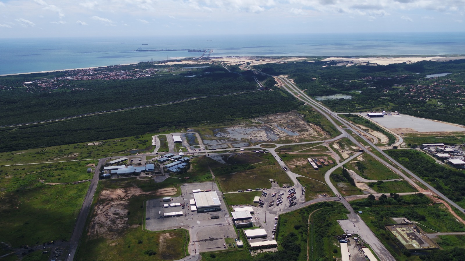Em primeiro plano aparecem áreas industriais, galpões e vias de acesso. Ao fundo, é possível ver o porto avançando sobre o mar, cercado por dunas e vegetação litorânea. A imagem destaca a integração entre a infraestrutura logística do complexo, a malha rodoviária e a costa cearense.