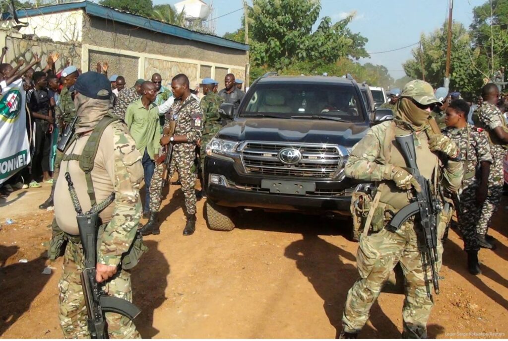 Homens armados e com os rostos cobertos fazem segurança junto a um veículo preto, cercados por militares e civis.