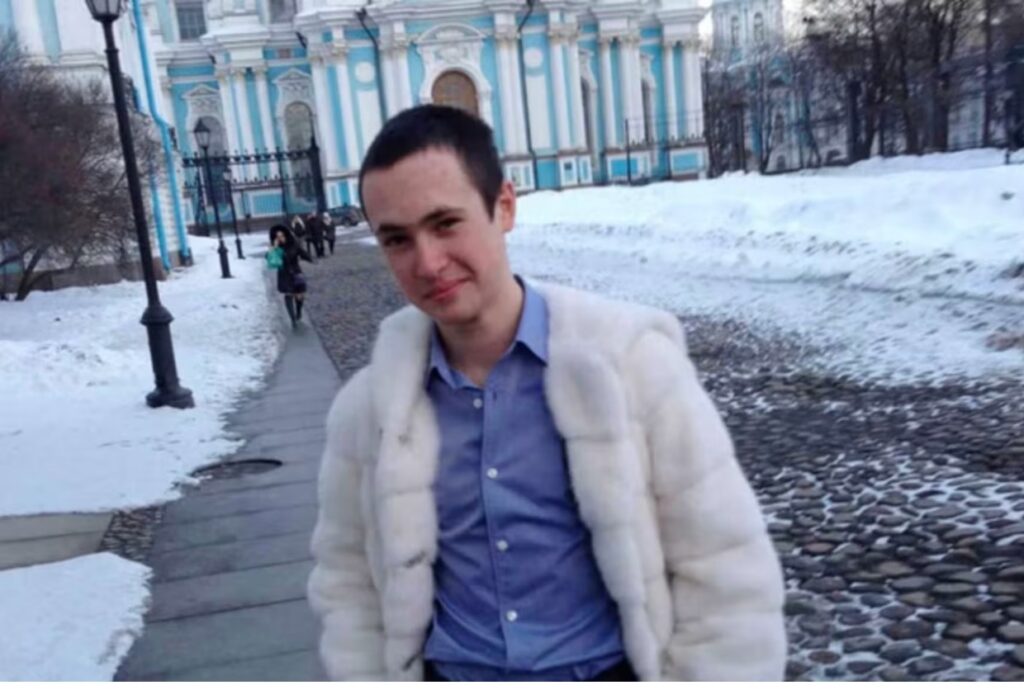 Pavel Prigozhin veste um casaco claro e camisa azul diante de um edifício, com neve ao redor.