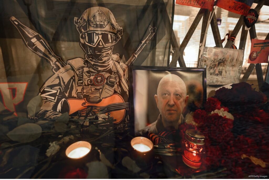 Retrato emoldurado de Yevgeny Prigozhin cercado por flores e velas, diante de uma bandeira com símbolos do Wagner.